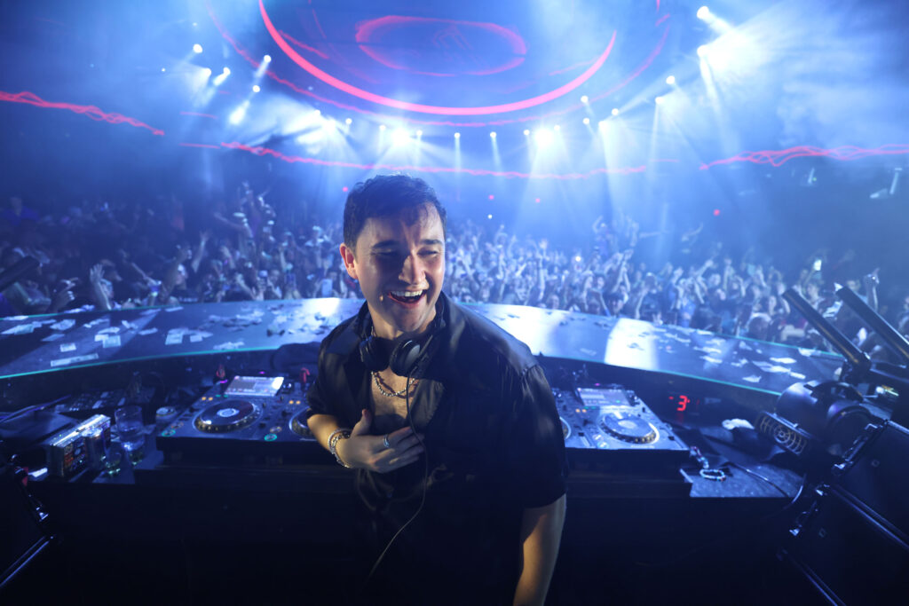 Up close shot at DJ booth of John Summit smiling with crowd cheering behind him.