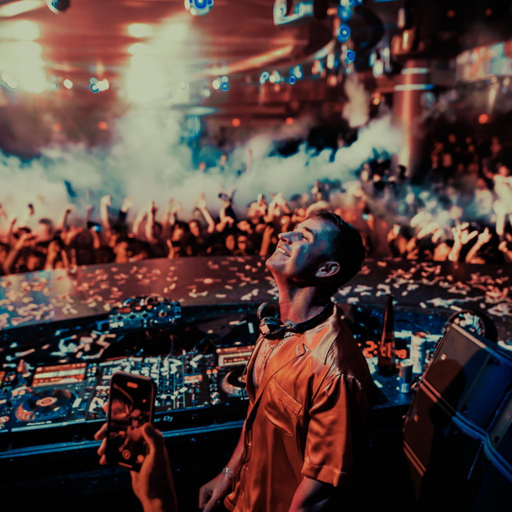 John Summit in DJ booth smiling with his eyes closed looking up with crowd and cryo behind him 