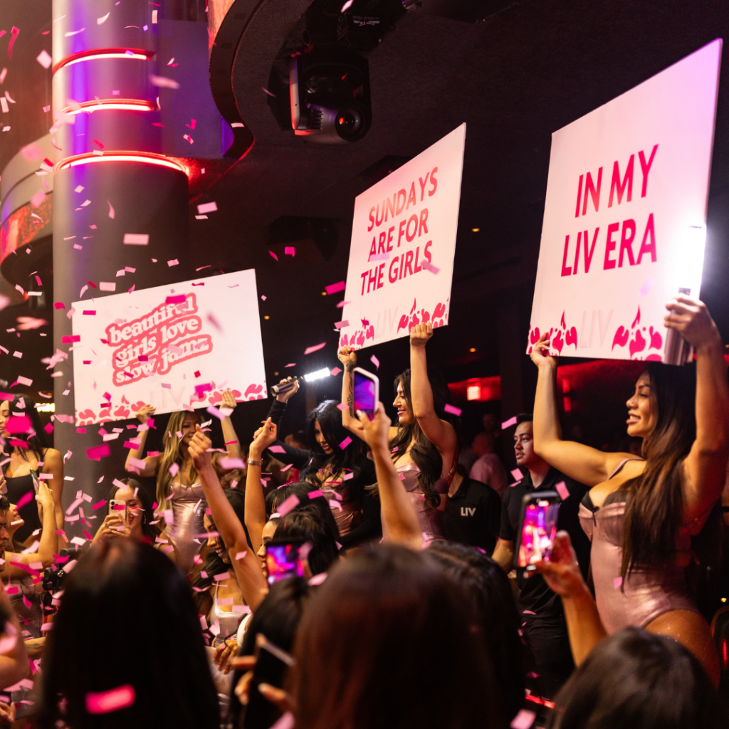 Three servers each holding a pink sign that says, "beautiful girls love slow jamz", "SUNDAYS ARE FOR THE GIRLS", and "IN MY LIV ERA" with pink confetti in the air