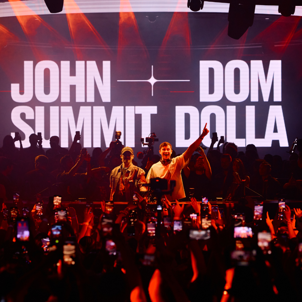 Shot from the crowd of Dom Dolla (left) and John Summit (right) performing in the DJ booth with their names on the LED screen behind them