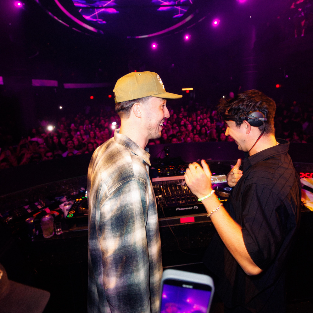 Cloonee (left) and John Summit (right) behind the DJ booth smiling and talking to each other with crowd behind them